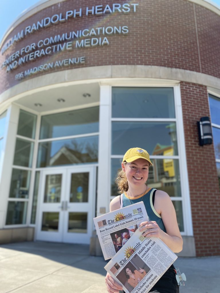 A female student stands in front of a campus building. She is wearing a yellow baseball cap and holding two copies of the student newspaper