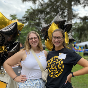Two women stand outside on the Saint Rose campus. They are smiling in front of black and gold balloons.